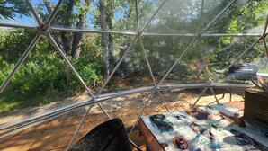 A geodesic dome structure with a view of a forested area through the transparent walls. Inside, there is a table with various items and a wicker basket.