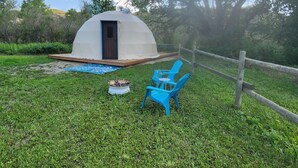 A dome-shaped tent with a door, a fire pit, and two blue chairs in a grassy area with a wooden fence. - #D3 Rainforest Getaway Dome- 2 Dbl beds-No pets Please (Drumheller)
