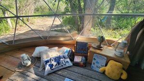 A cozy, rustic cabin with a large window overlooking a forested area. Inside, there is a wooden table with a globe, a lantern, and a plush toy. - #D4 Badlands Bunker Dome-1 Queen bed -Pet friendly (Drumheller)