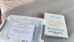 A welcome package on a bed with a patterned pillow and a grey blanket. - #D4 Badlands Bunker Dome-1 Queen bed -Pet friendly (Drumheller)