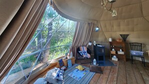 A cozy room with a large window, wooden flooring, a small table with books and a globe, and a chair with a blanket. - #D4 Badlands Bunker Dome-1 Queen bed -Pet friendly (Drumheller)