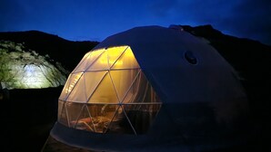 Een geodetische koepelstructuur met warm binnenlicht, gelegen op een houten terras met een bergachtige achtergrond.