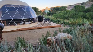 Een geodetische koepeltent met een houten terras en een stoel, omgeven door natuurlijke begroeiing en heuvels op de achtergrond.