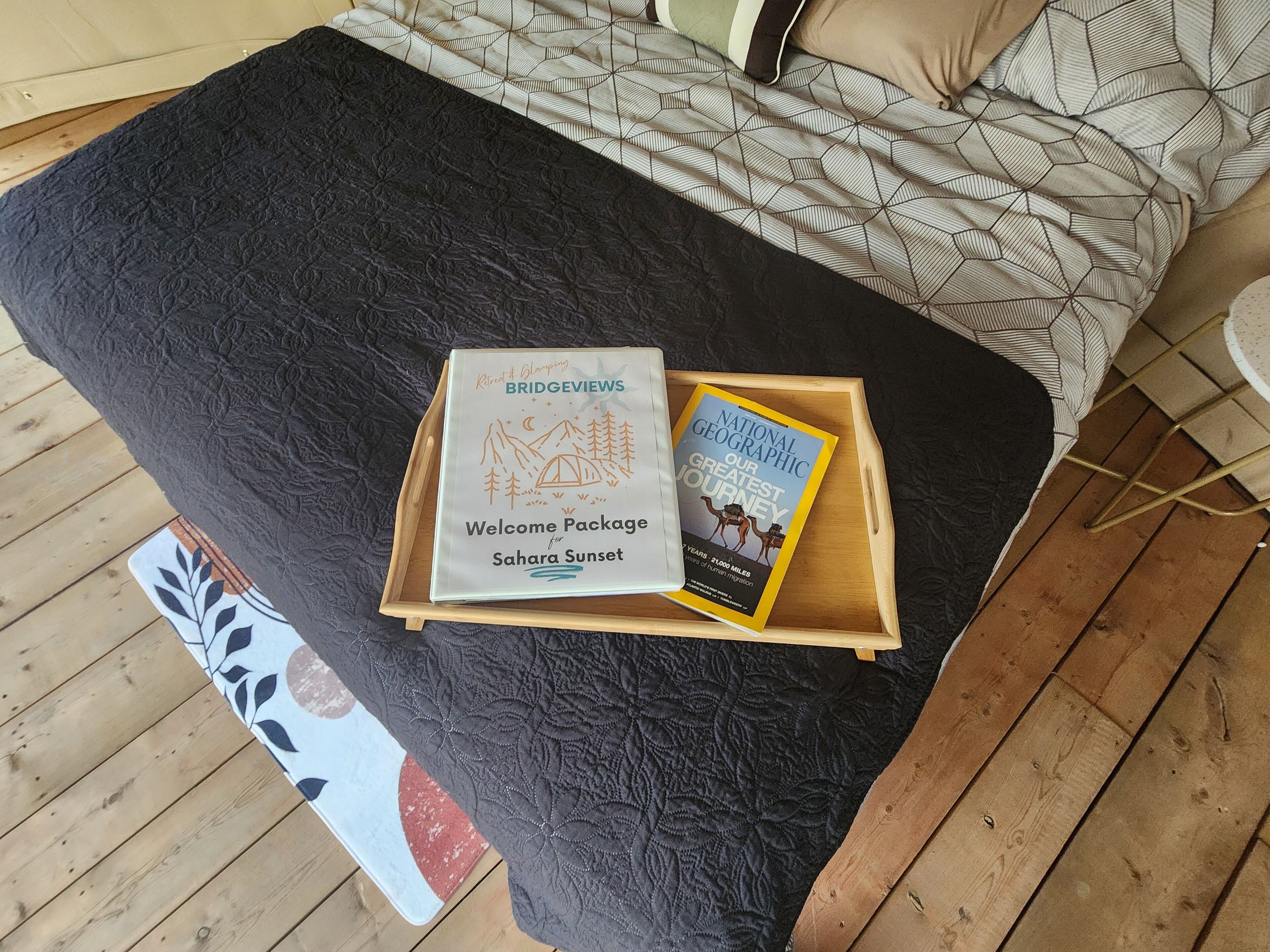 A bed with a patterned bedspread, a tray with a welcome package and a book titled "National Geographic Our Greatest Journey," and a wooden floor.