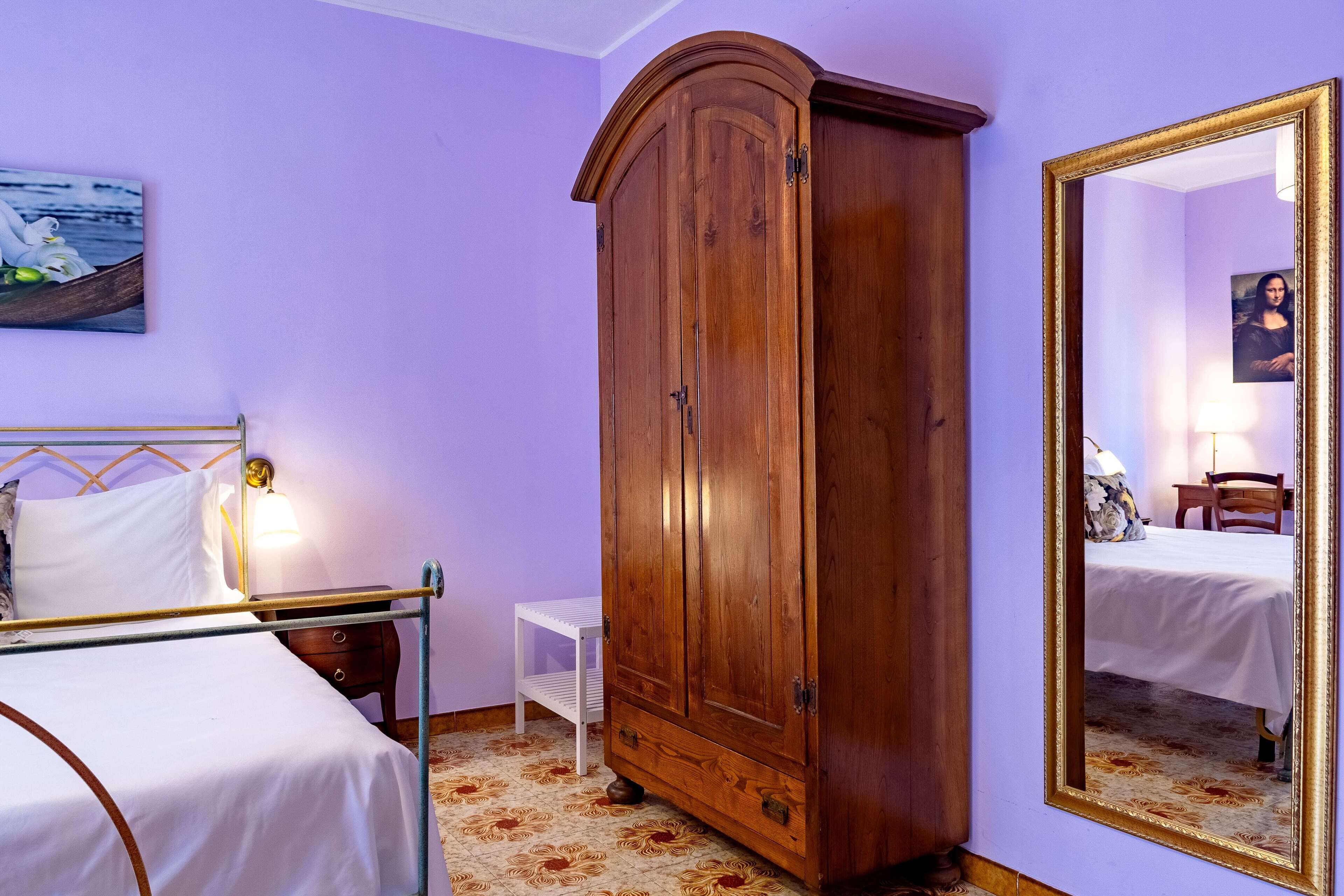 Um quarto com cama, guarda-roupa de madeira, mesinhas de cabeceira pequenas e brancas, espelho e um quadro na parede.