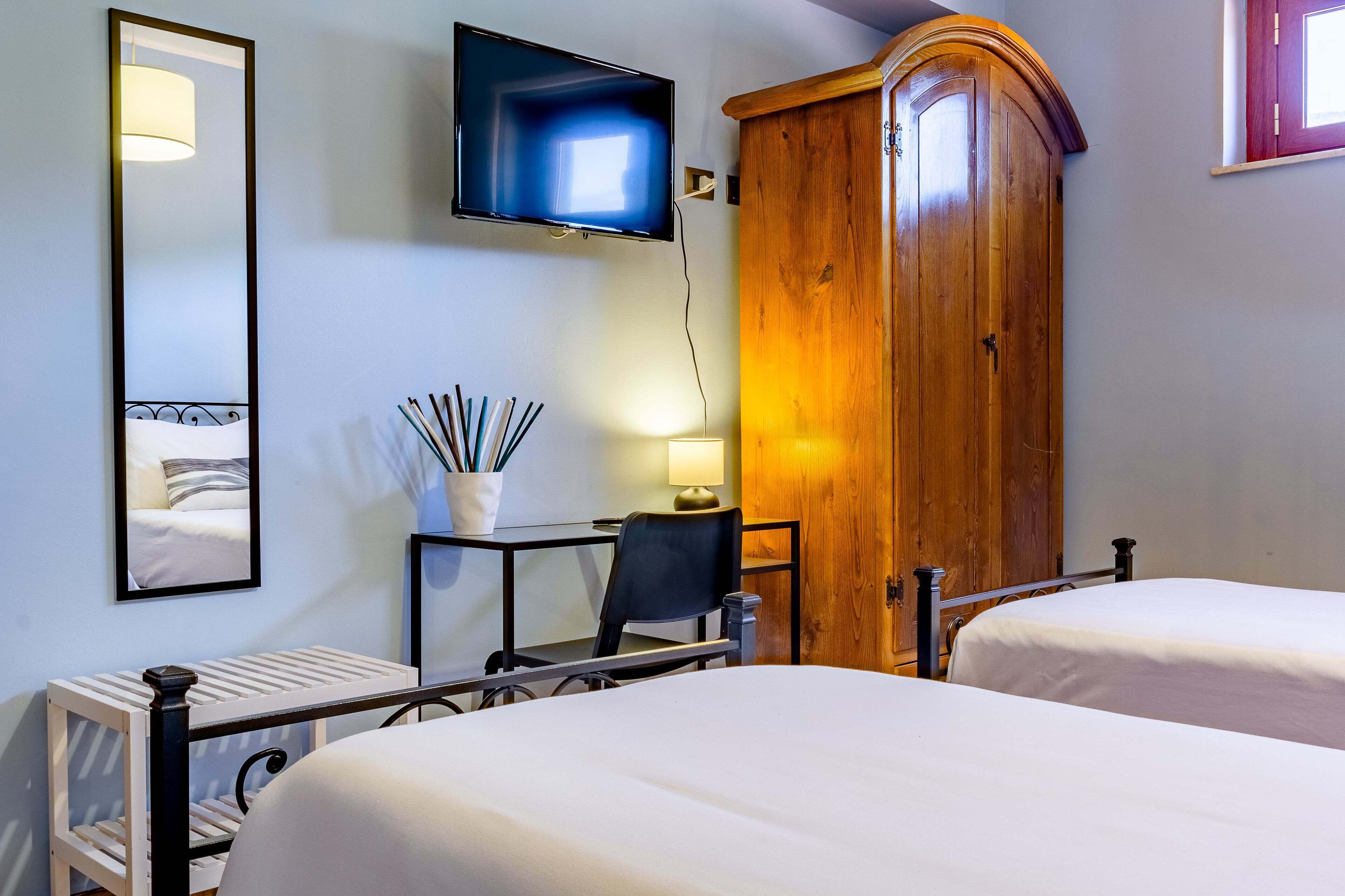 Habitación de hotel con dos camas, un televisor montado en la pared, un armario de madera, un escritorio con una silla y una mesita de noche con una lámpara.