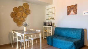 A modern living room with a teal sofa, a dining table with white chairs, a wall-mounted shelf with kitchen appliances, and decorative woven wall art. - Open Horizon - Short Rent (Zafferana Etnea)