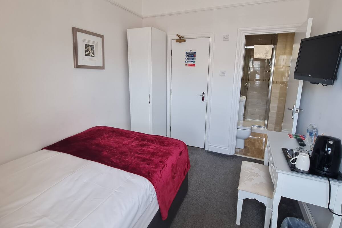 Habitación de hotel con cama, escritorio con cafetera, televisor, armario y baño visible a través de una puerta abierta.