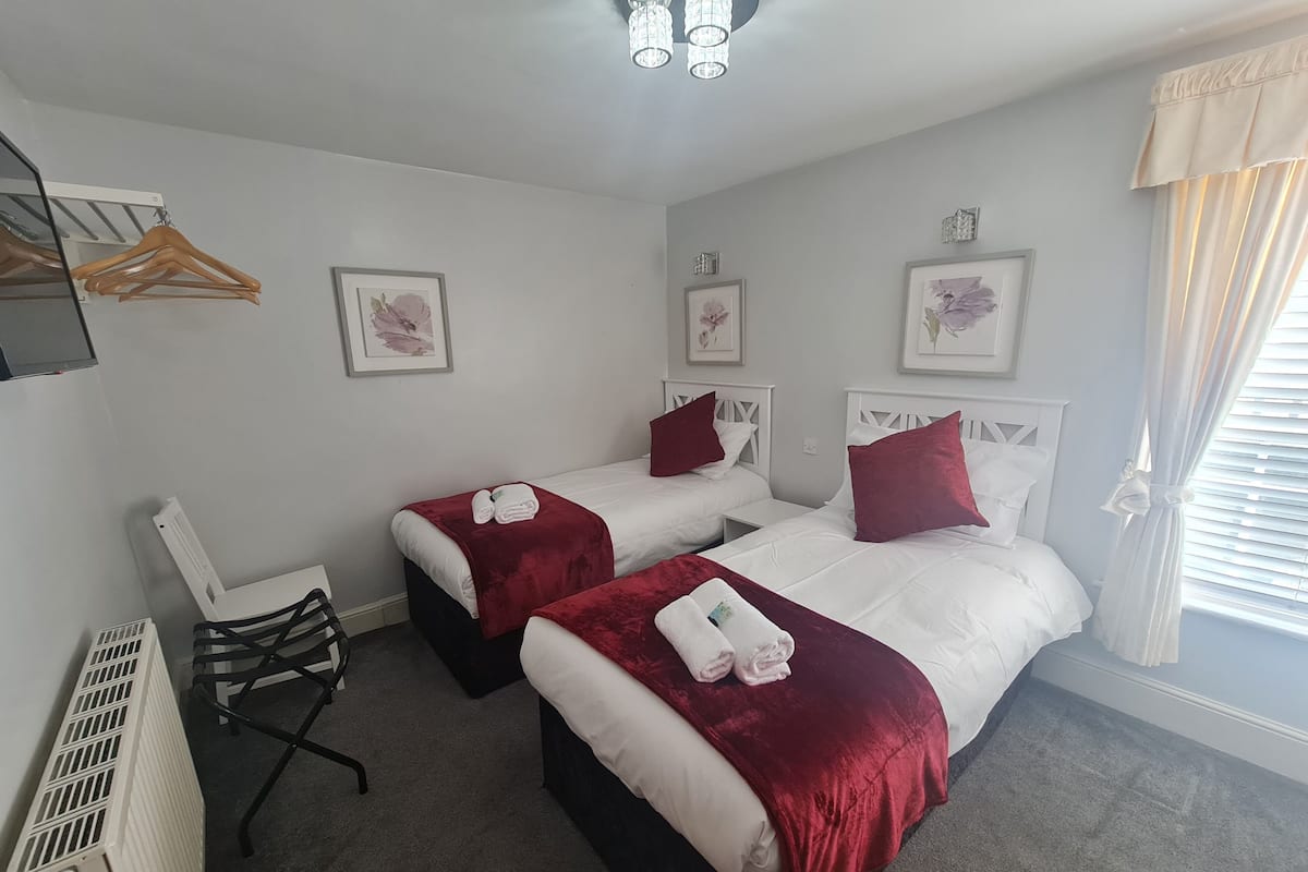 Habitación de hotel con dos camas, cada una con ropa blanca y cojines rojos. Se observan cuadros en las paredes, un radiador y una silla.