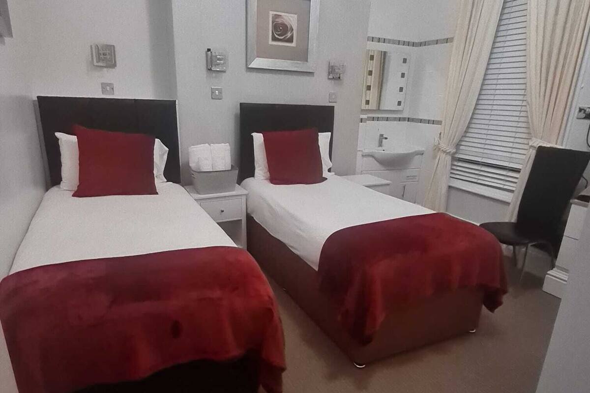 Una habitación de hotel con dos camas, cada una con una manta roja, una mesita de noche con lámpara y un pequeño cuadro en la pared.