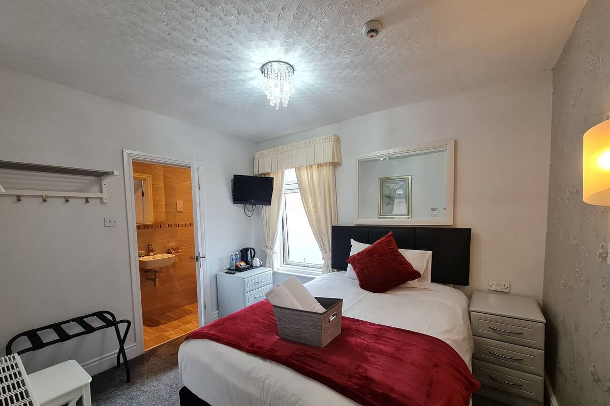 Habitación de hotel con cama, mesita de noche, escritorio, espejo, televisor y baño a la vista a través de una puerta abierta.