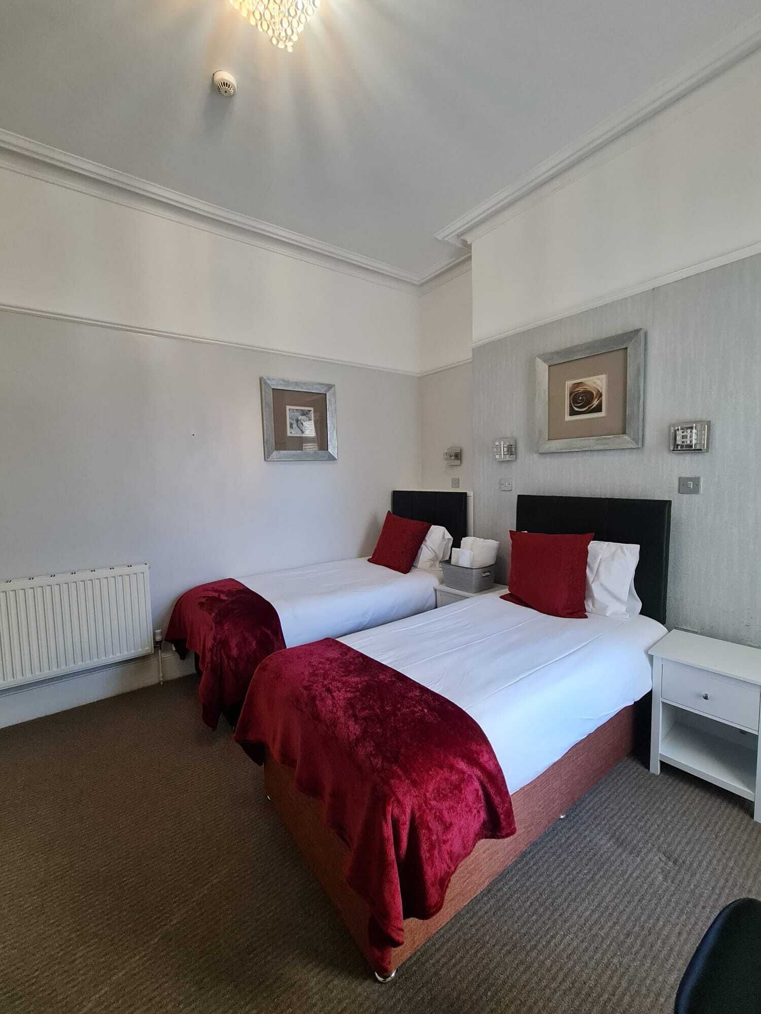 Habitación de hotel con dos camas, ropa de cama blanca, almohadones rojos y un radiador. Se observan también una mesita de noche y un aparador blancos.