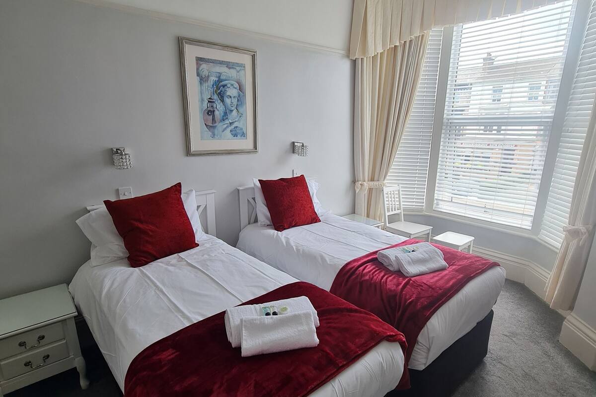 Habitación de hotel con dos camas, cada una con ropa de cama blanca y detalles rojos, una obra de arte enmarcada sobre las camas, una mesita de noche con lámpara y una ventana con persianas.