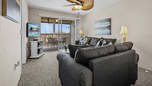 Ein Wohnzimmer mit grauem Sofa, Deckenventilator, Fernseher und einem Balkon mit Tisch und Stühlen.