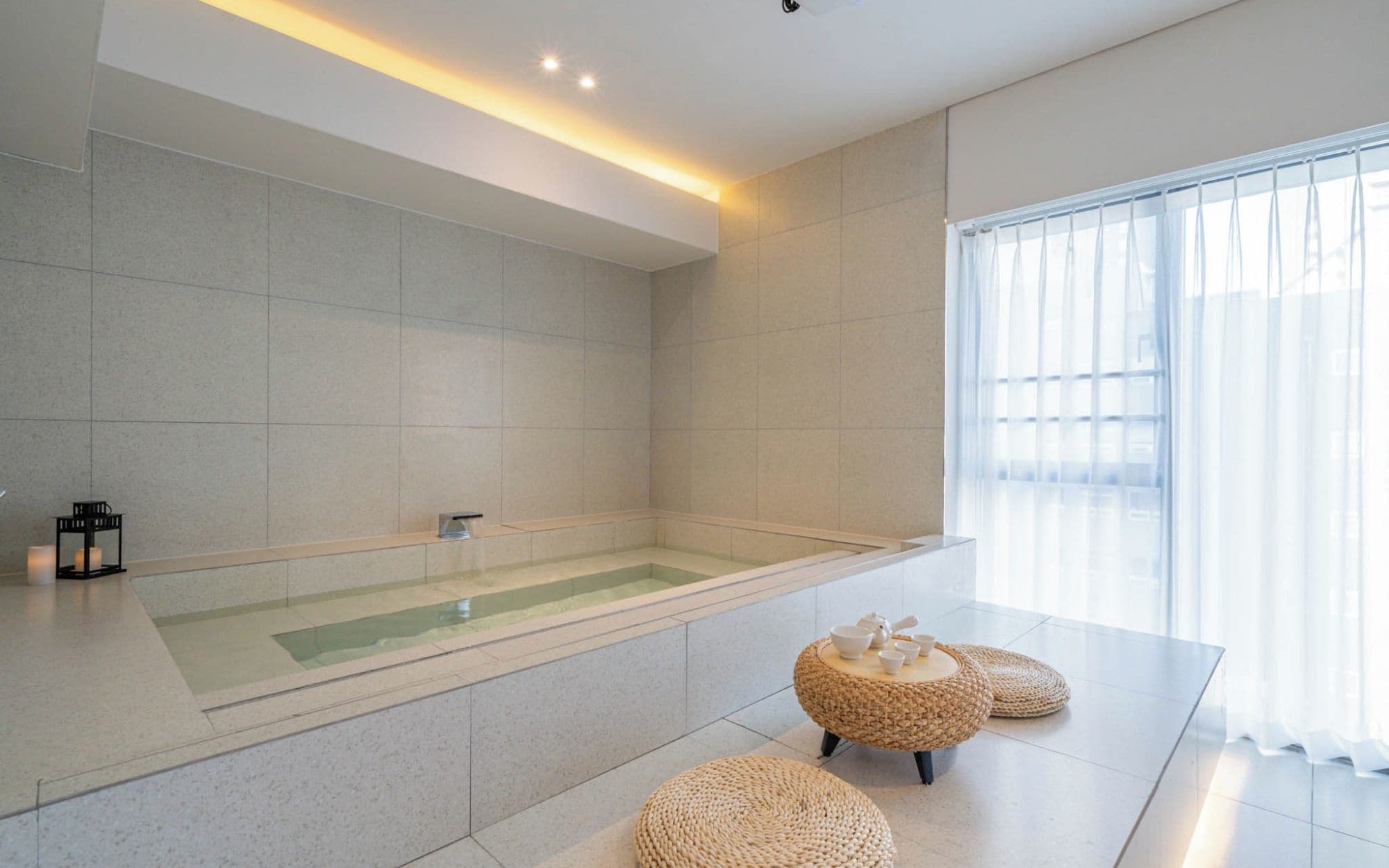 A modern bathroom with a large bathtub, a glass-enclosed shower, and a seating area with woven stools.