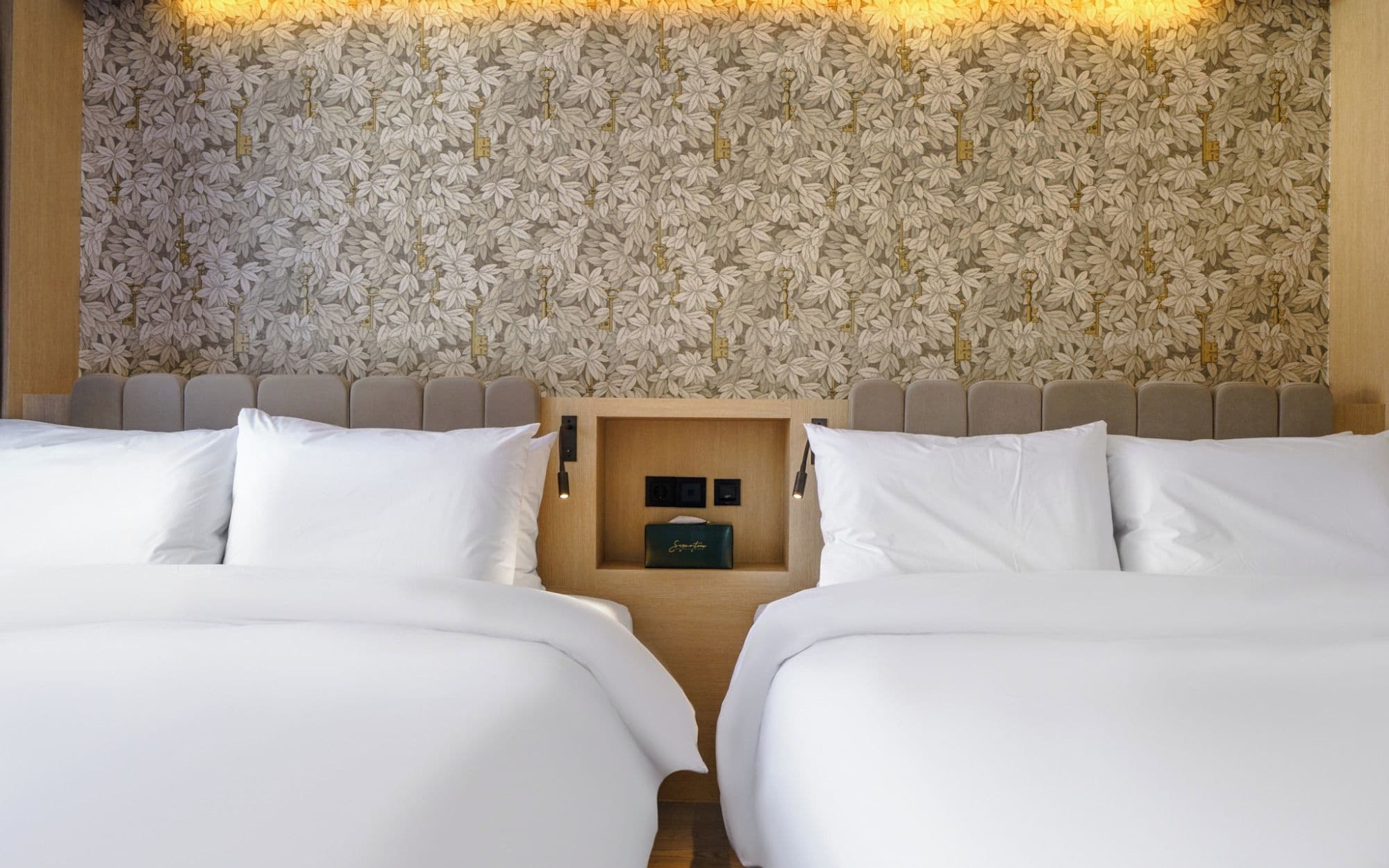 Een hotelkamer met bloemenbehang, een houten hoofdbord en een inbouwkastje met een klein zwart voorwerp.
