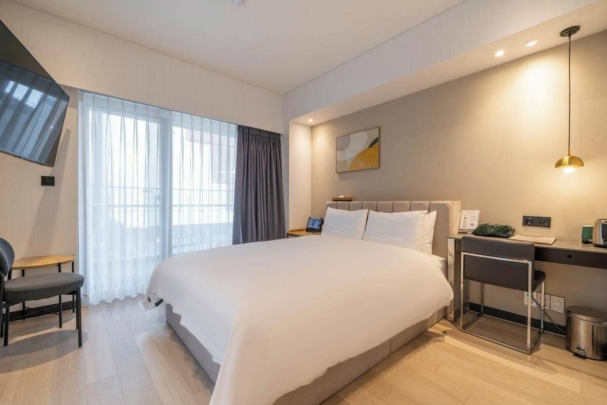 Ein modernes Hotelzimmer mit einem großen Bett, einem Schreibtisch, einem Sessel, einem Fernseher und einem Fenster mit Vorhängen.