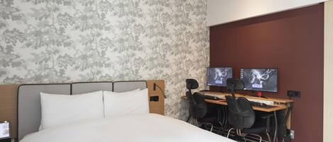 A hotel room with a bed, two monitors, a desk, and a chair.