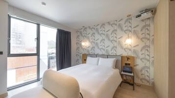 A hotel room with a large bed, a desk with a laptop, a projector, and a view of the city through a large window.