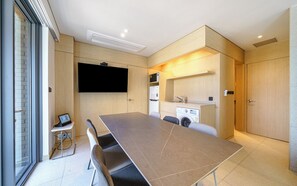 A modern hotel room with a large dining table, a flat-screen TV mounted on the wall, a small kitchenette, and a sliding door leading to a balcony. - Brown-dot Hotel Haeundae (Busan)