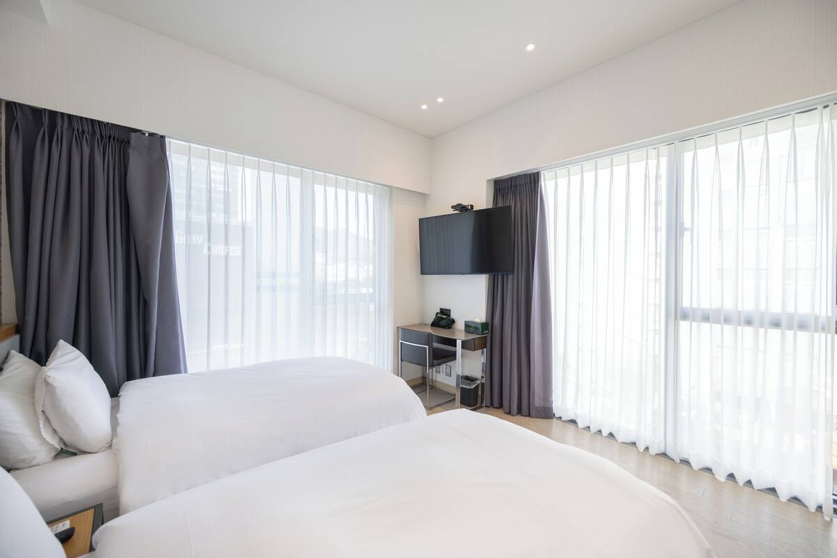 Ein Hotelzimmer mit zwei Betten, einem Schreibtisch, einem Fernseher und großen Fenstern mit Vorhängen.
