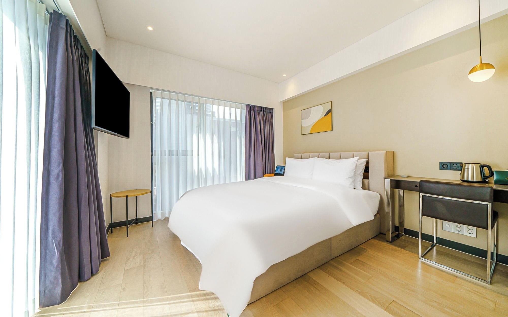 Habitación de hotel con una cama grande, un televisor de pantalla plana montado en la pared, una mesita de noche con una lámpara y una cafetera.