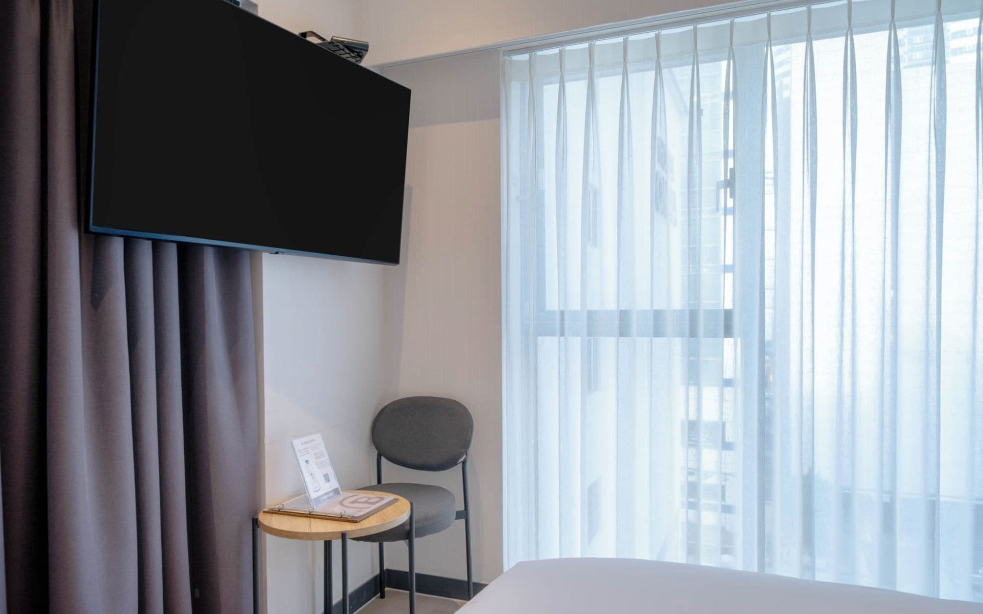 A hotel room with a bed, a desk with a chair, a flat-screen TV mounted on the wall, and a window with sheer curtains.
