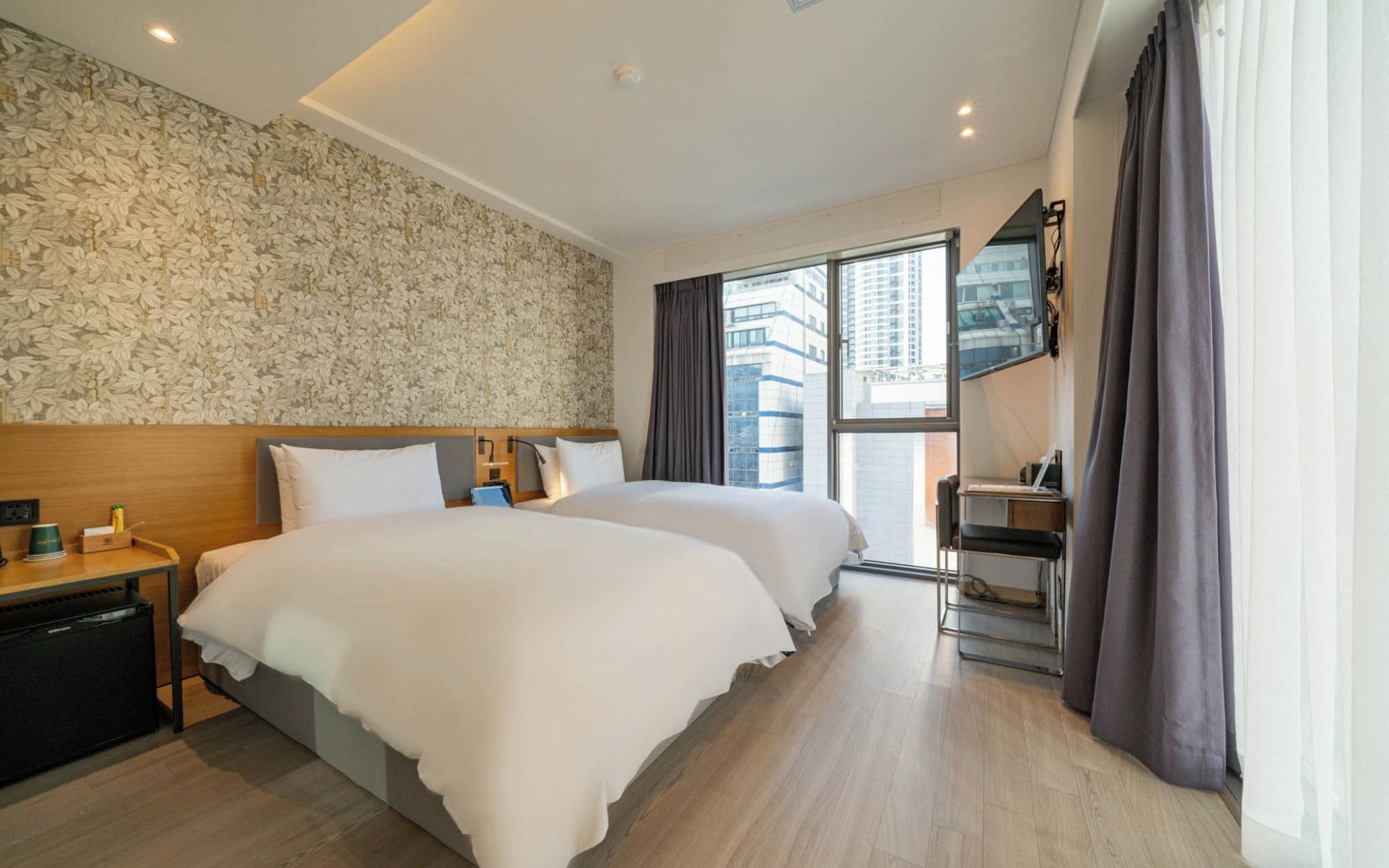 A hotel room with two beds, a desk, a chair, a window with a view of buildings, and a patterned wall.