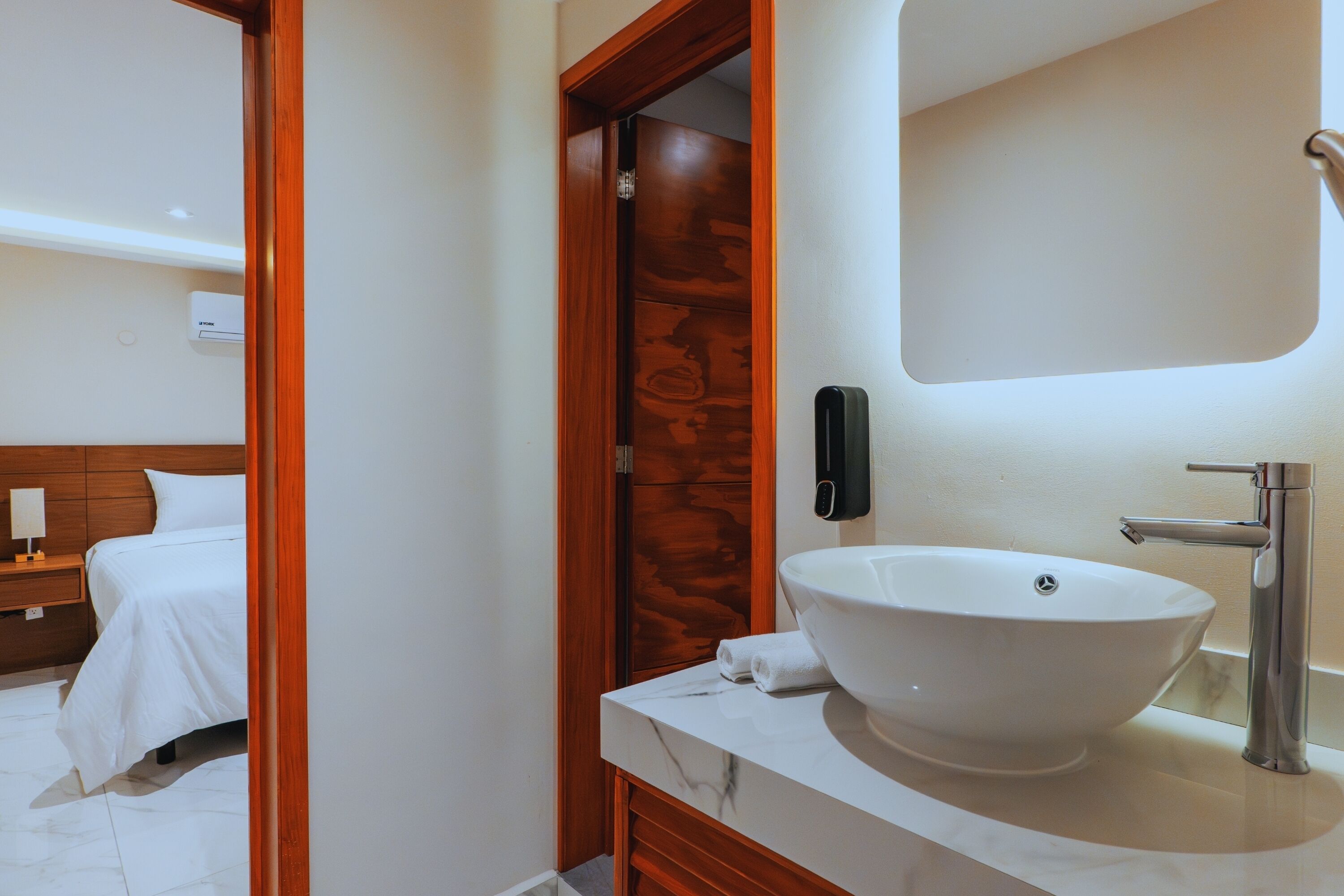 A modern bathroom with a white marble sink, a wooden door, and a visible bed in the adjacent room.