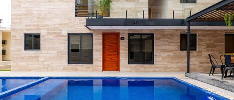 A modern building with a swimming pool, a wooden door, and a balcony with plants.