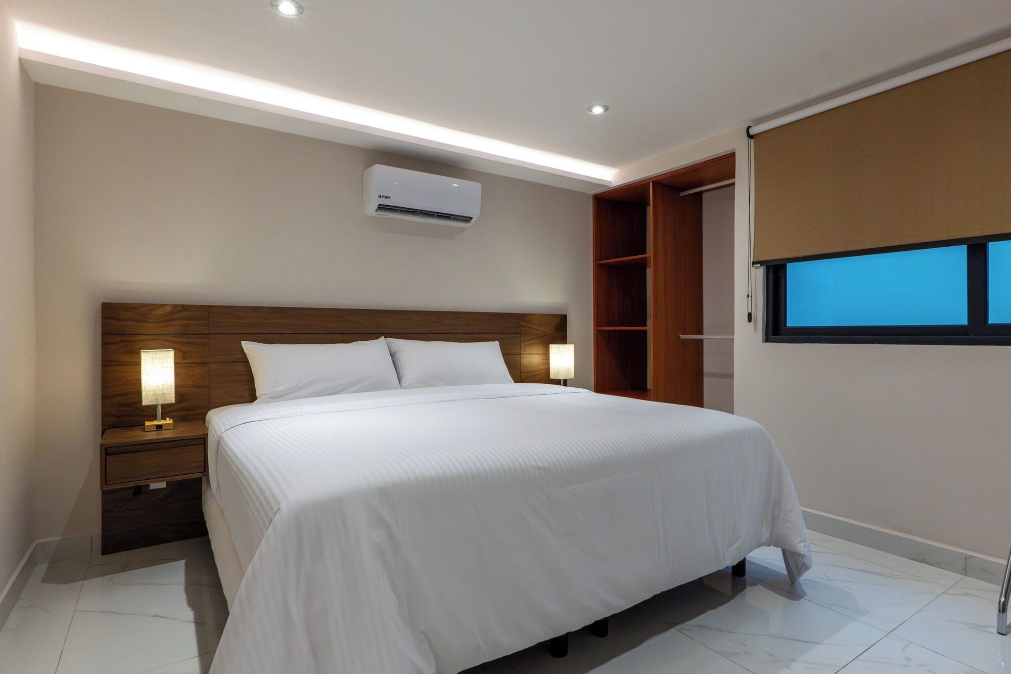 A modern hotel room with a large bed, wooden headboard, bedside lamps, an air conditioning unit, a wardrobe, and a window with a view.