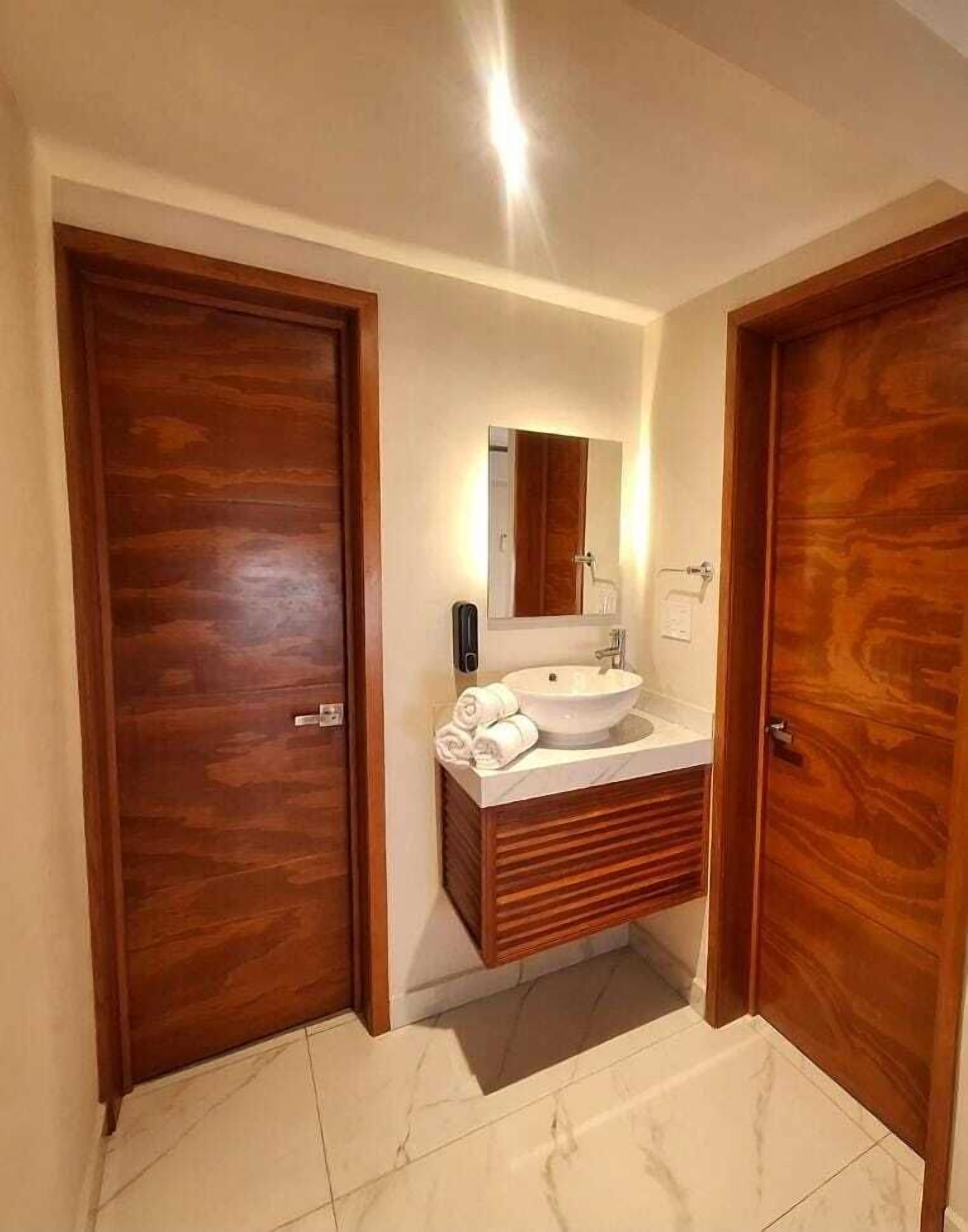 Casa de banho com porta de madeira, pia branca, espelho e porta-toalhas.