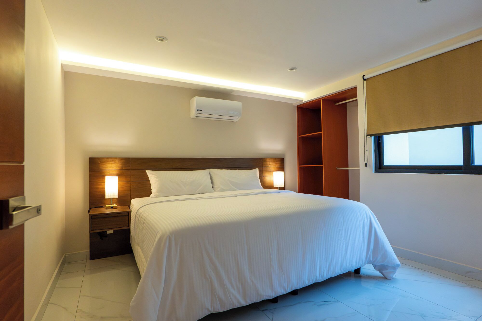 A hotel room with a bed, nightstands, a wall-mounted air conditioner, a window with a blind, and a wooden wardrobe.