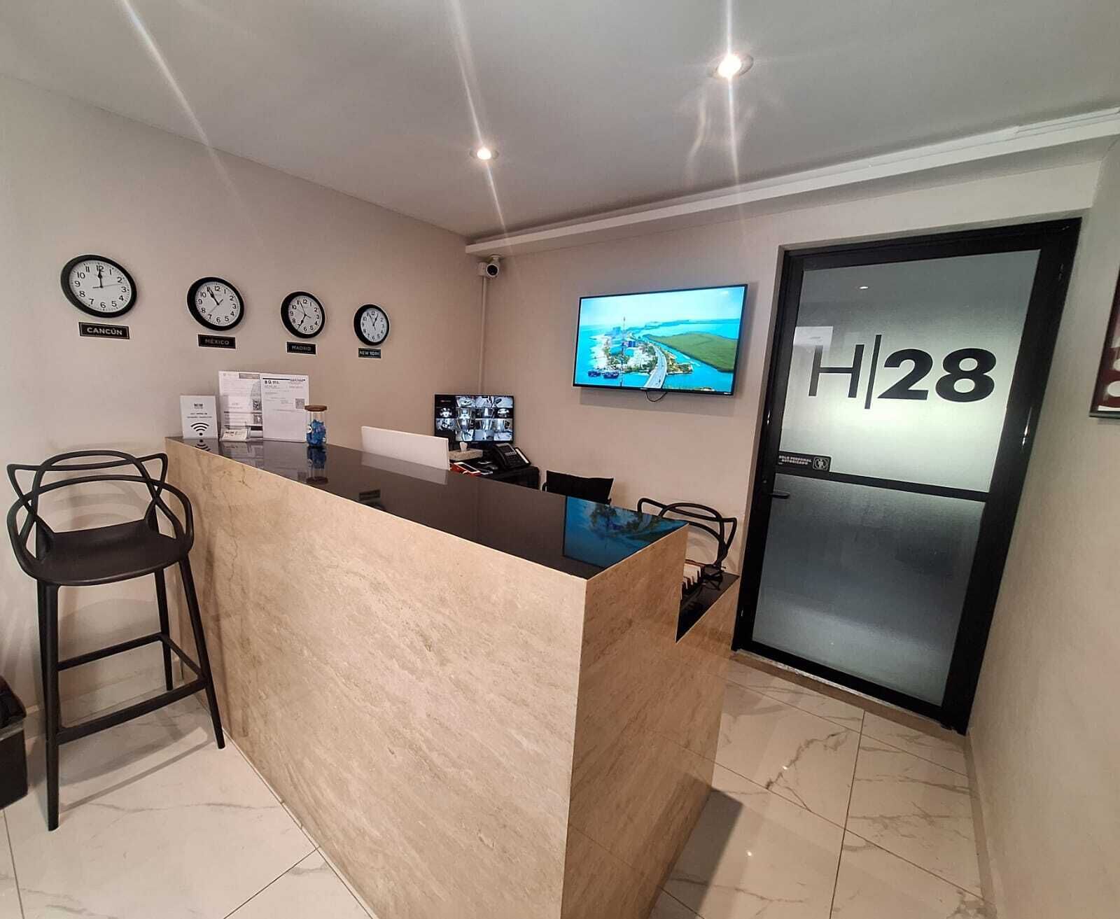 A hotel reception area with a reception desk, chairs, clocks, a television, and a door marked 'H|28'.