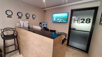 A hotel reception area with a reception desk, chairs, clocks, a television, and a door marked 'H|28'.