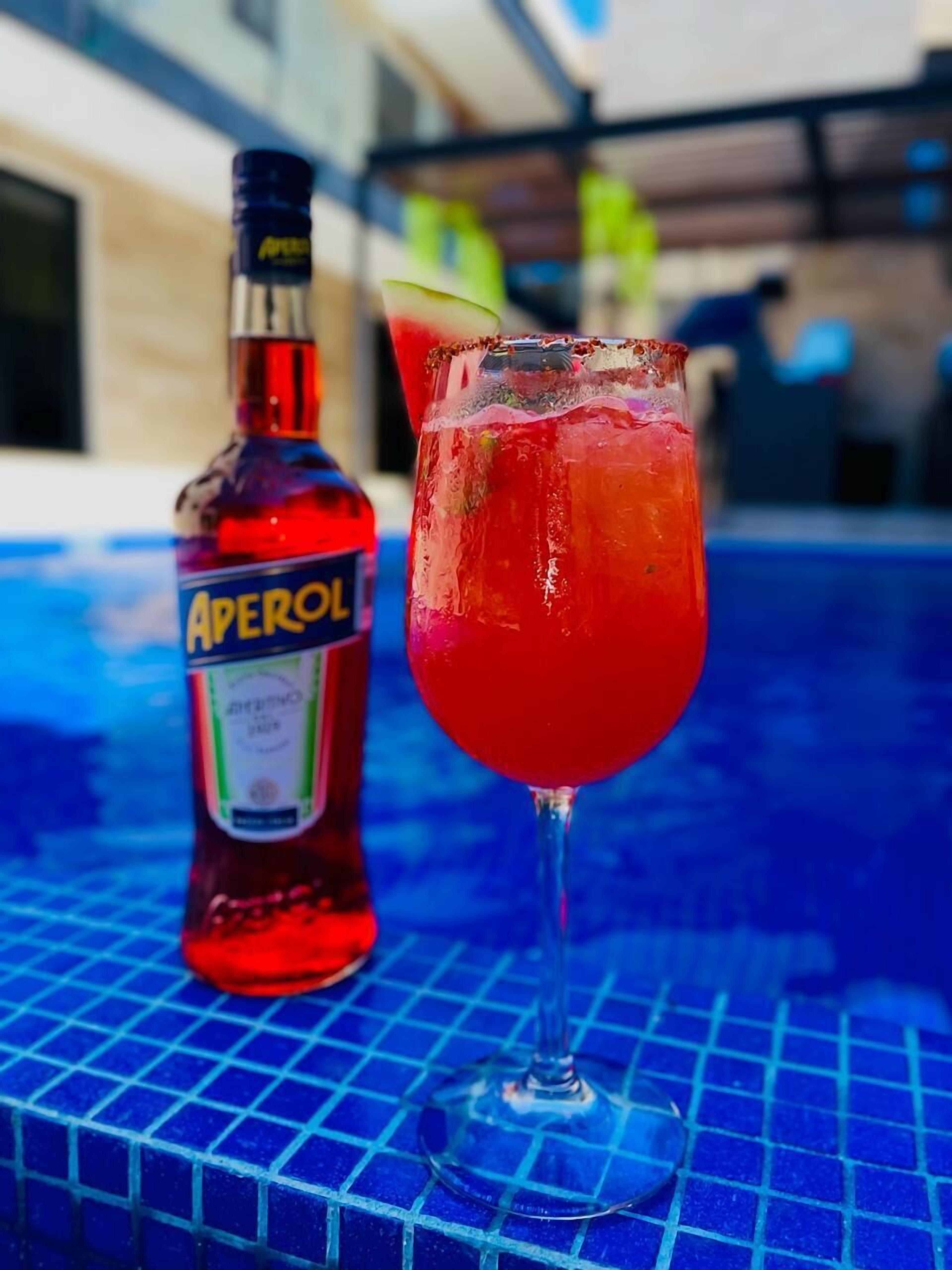 A bottle of Aperol and a glass of red cocktail on a blue tiled surface.
