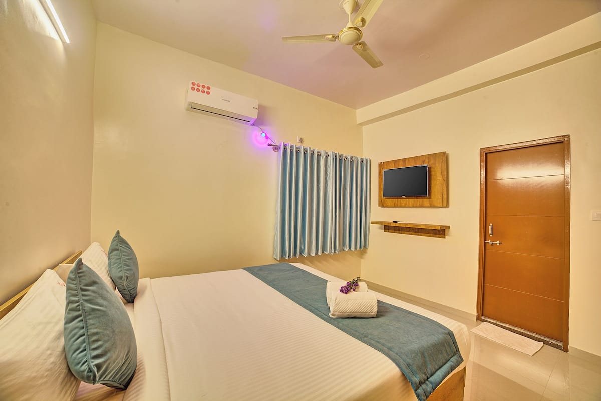 Une chambre d’hôtel comprenant un lit, des oreillers, une table de chevet, un téléviseur, un appareil de climatisation, un ventilateur de plafond et une porte.