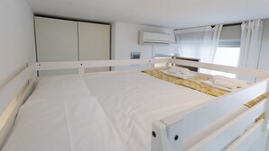 A single bed with white bedding, a bedside table, a wardrobe, an air conditioning unit, and a window with curtains. - GuestHost - Comfortable 28 sqm studio apartment, ideal for 2 people, located on the first floor of a renovated building with elevator (3 stairs to access it). The accommodation is located in a quiet residential area, about 50 meters from the beach.A 2-mi (Rimini)