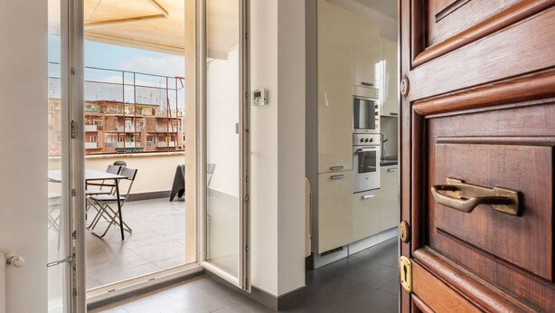 An open sliding glass door leading to a balcony with a table and chairs, a kitchen with cabinets and appliances, and a wooden door with a brass handle. - GuestHost - Trastevere Station Bright Apartment with Terrace (Roma)