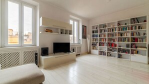 A spacious living room with a flat-screen TV, a bookshelf, and a large window. - GuestHost - Trastevere Station Bright Apartment with Terrace (Roma)