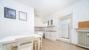 Une cuisine avec des meubles blancs, une table à manger avec des chaises, un tableau d’affichage en liège accroché au mur et un radiateur.