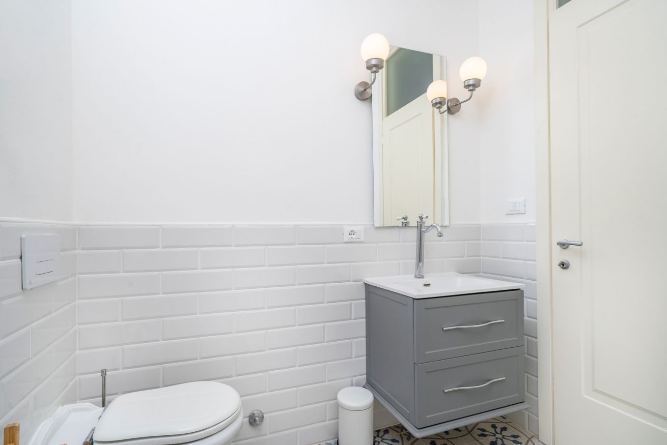 Et badeværelse med hvide flisemure, et hvidt toilet, et gråt skab med skuffer og et spejl med vægmonteret lys.