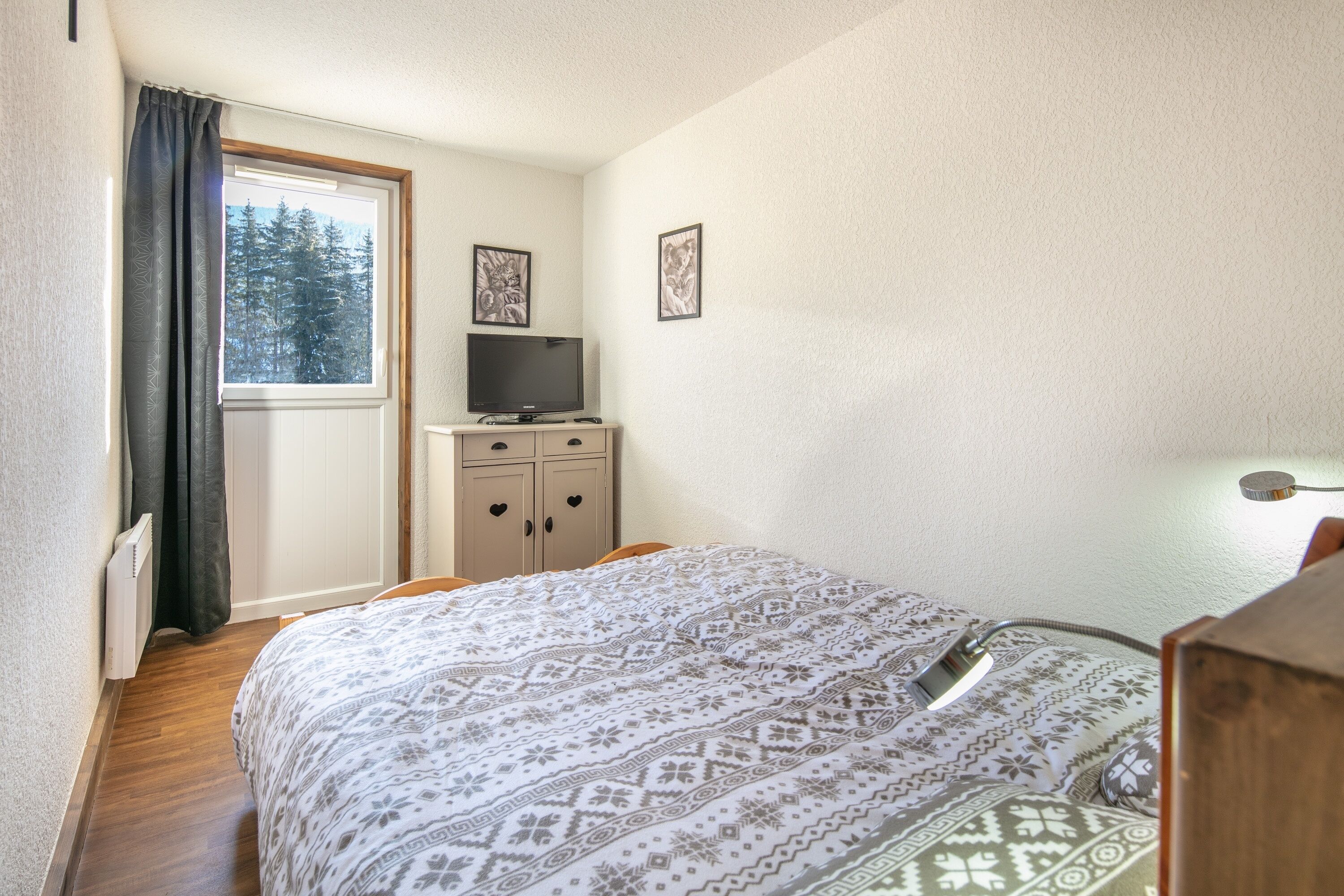 Een slaapkamer met een bed, een dressoir, een televisie en een raam met uitzicht op bomen.