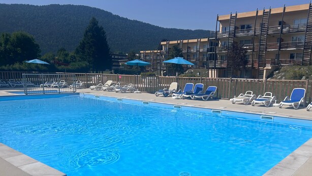 Ein Schwimmbecken mit Liegestühlen und Sonnenschirmen, umgeben von einem Holzzaun und Gebäuden mit Balkonen.
