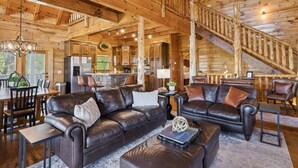 A spacious log cabin interior with a dining area, leather sofas, a staircase, and a kitchen. - Falling Leaf Hideaway – Alpine Mountain Village Cabin with Hot Tub, Theater, Game Room, Pools! (Pigeon Forge)
