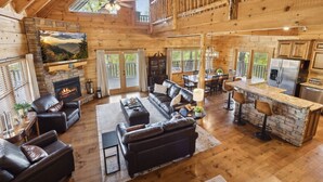 A spacious log cabin interior with a fireplace, leather sofas, a dining area, and a kitchen. - Falling Leaf Hideaway – Alpine Mountain Village Cabin with Hot Tub, Theater, Game Room, Pools! (Pigeon Forge)