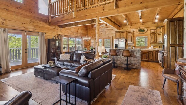 A spacious living room with a large black leather sofa, a dining area, and a kitchen with stone countertops. - Falling Leaf Hideaway – Alpine Mountain Village Cabin with Hot Tub, Theater, Game Room, Pools! (Pigeon Forge)
