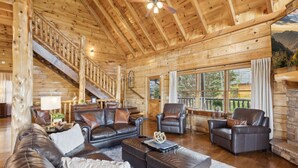 A spacious living room with a wooden interior, leather furniture, a staircase, and large windows offering a view of the outdoors. - Falling Leaf Hideaway – Alpine Mountain Village Cabin with Hot Tub, Theater, Game Room, Pools! (Pigeon Forge)