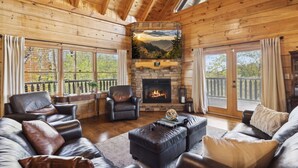 A cozy cabin interior with a stone fireplace, leather sofas, and a balcony with a view of the mountains. - Falling Leaf Hideaway – Alpine Mountain Village Cabin with Hot Tub, Theater, Game Room, Pools! (Pigeon Forge)