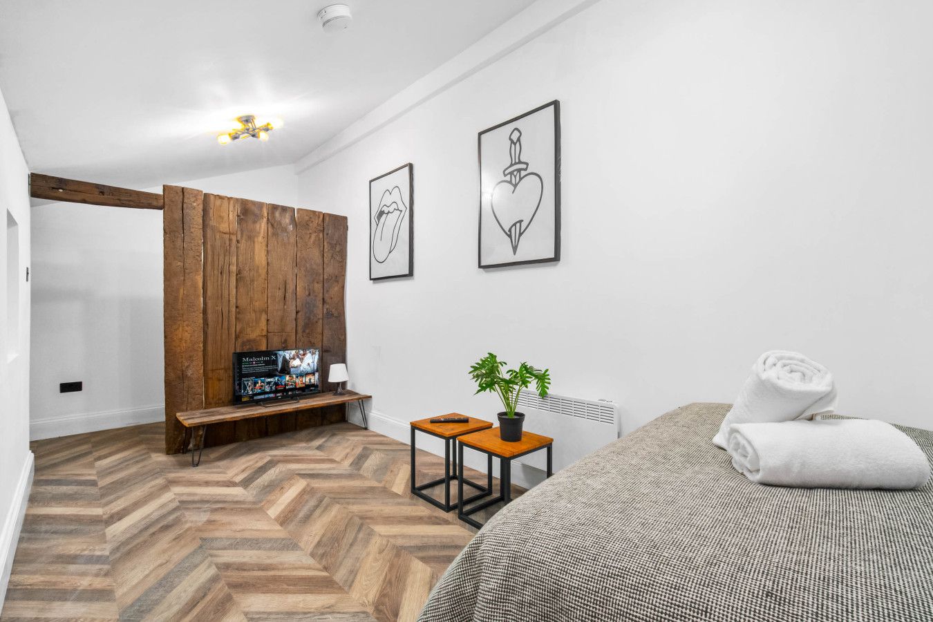 Un dormitorio moderno con piso de espiga, un banco de madera, un televisor, una mesita con una planta y una cama con una toalla enrollada.